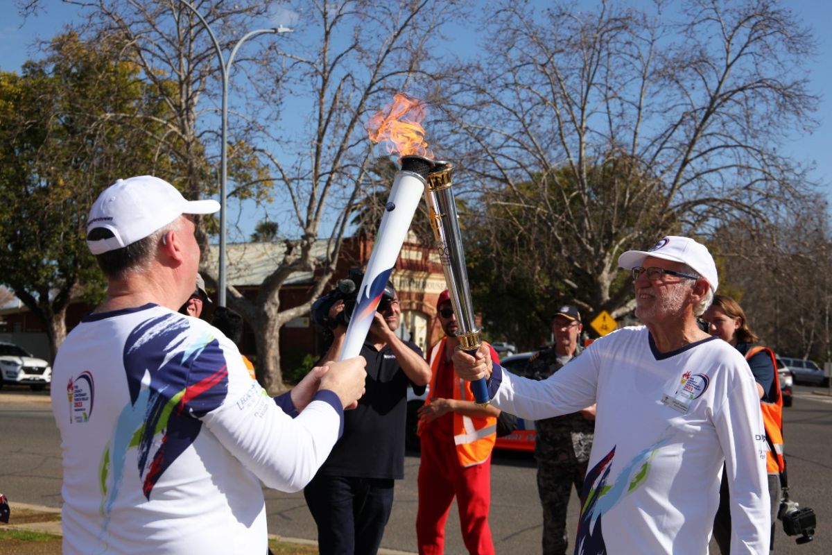 Two men in white shirts and shorts carry relay torches with flames, surrounded by media taking photos and video