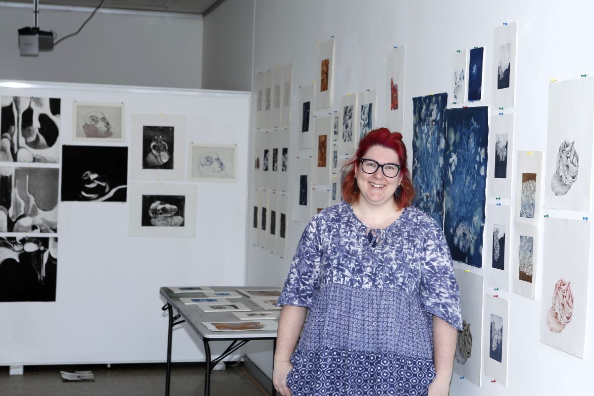 A woman wearing a blue and white dress stands in front of her artworks in a gallery space. 