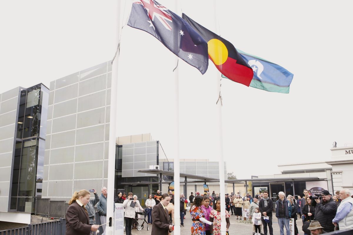 Young people raise the Australian, Aboriginal and Torres Strait Islander flags in front of a large crowd.