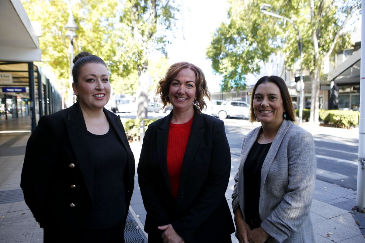 Three women dressed in business suits standing beside a main street with shops, trees and vehicles in the background.