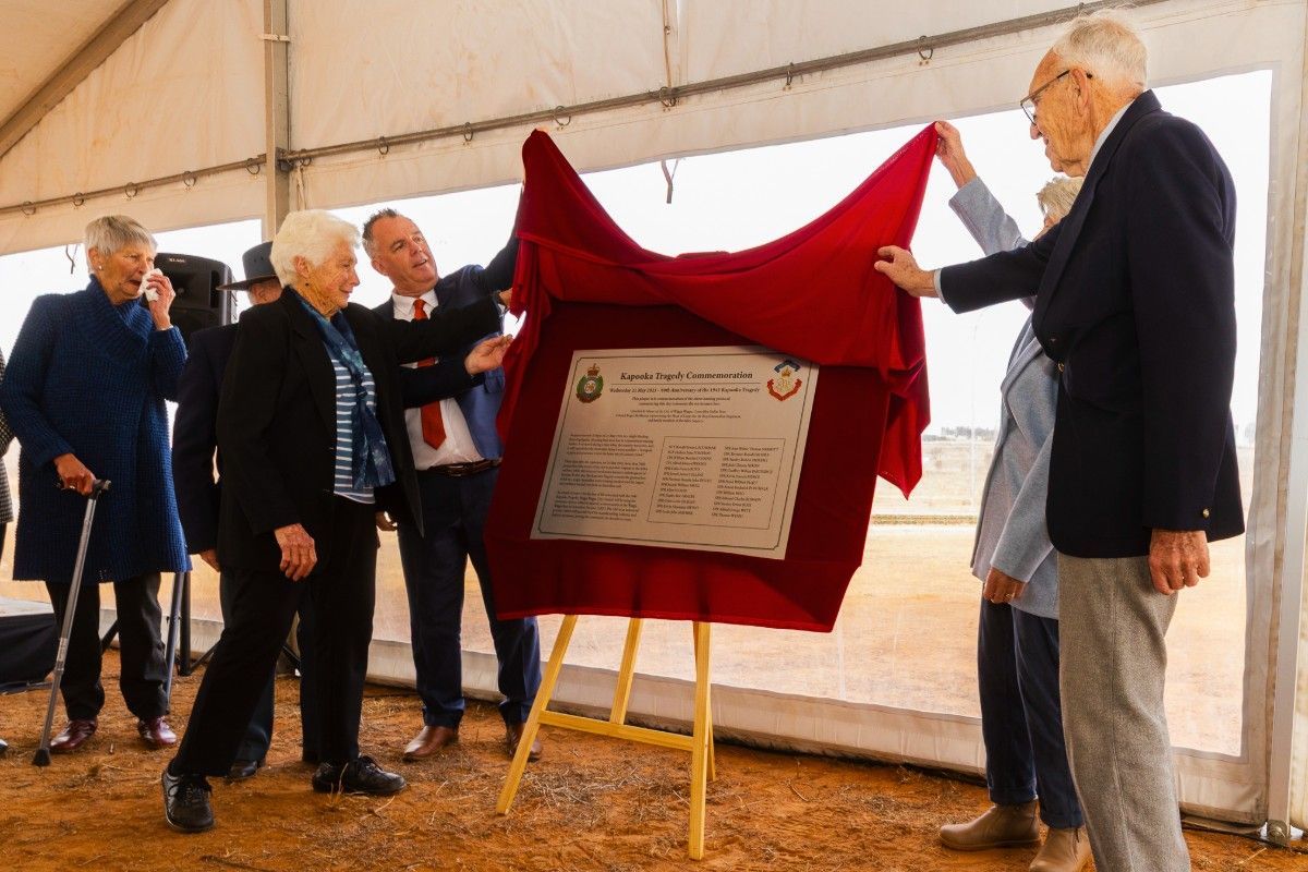 Three men and two women smile as they work together to lift up the edge of a red velvet curtain, in order to unveil a large silver memorial plaque.