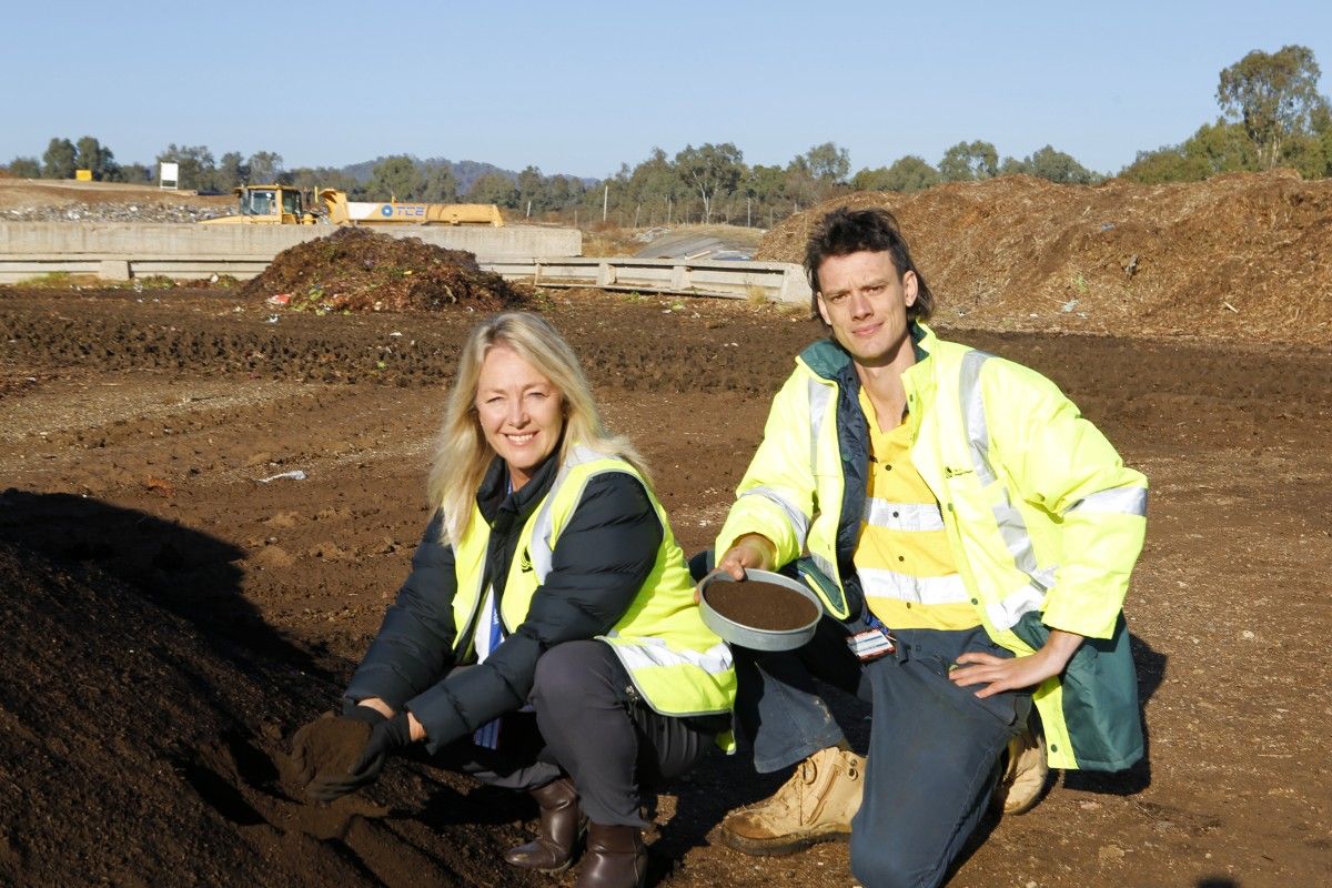 Woman and man in hi-visibility tops crouched beside a pile of compost, with the man holding a metal mesh strainer, or sieve, in his right hand.