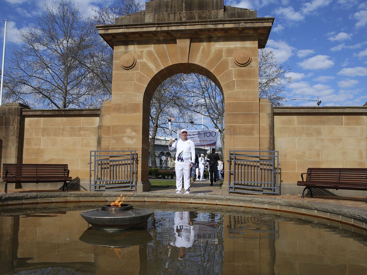 Wagga Mayor Councillor Dallas Tout walks under the Memorial Arch at Victory Memorial Gardens, with Eternal Flame in foreground and Defence personnel carrying the Legacy Centenary Torch Relay banner in the background