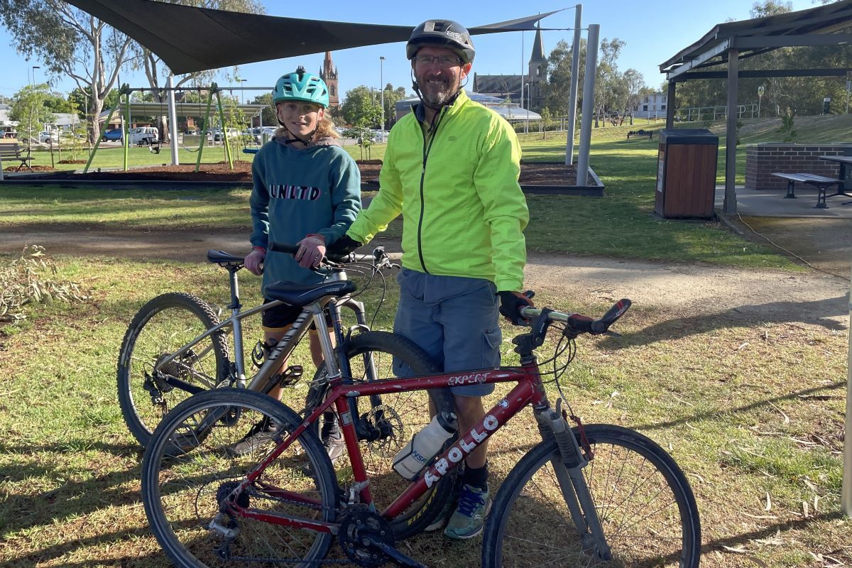 A man and a young person stand with their bicycles in a public park. 