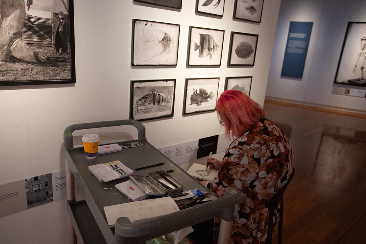 A woman sits in an art gallery drawing sketches, she is side on to the camera and there is a trolley with drawing implements next to her.