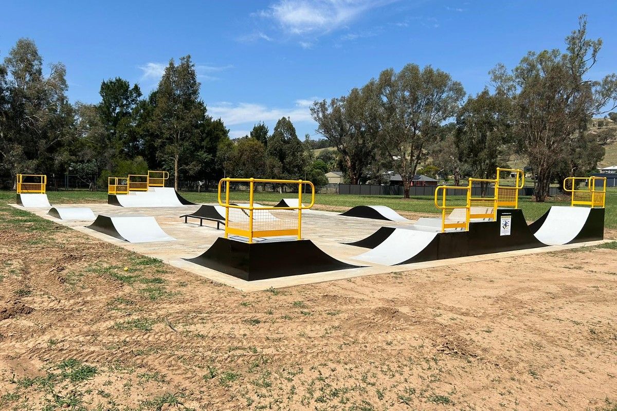 Newly constructed concrete and steel skatepark in open area, surrounded by grass and trees.
