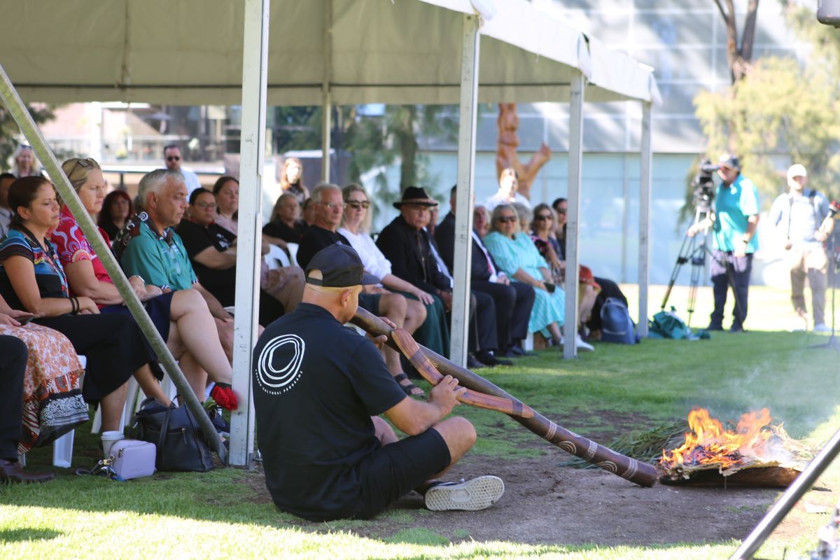 A man is sitting on the ground in front of a seated audience, he is playing the didgeridoo next to a coolamon with leaves that are on fire. 