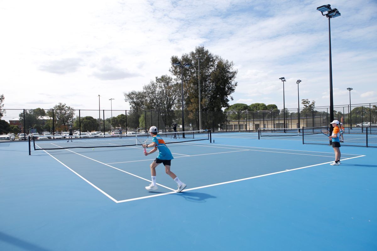 Two young people in brightly coloured sports gear stand closest to the camera while two are in the background. All four are working together to play doubles tennis on a blue tennis court.