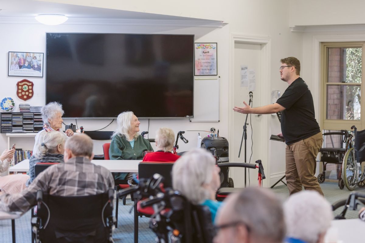 A man in an Opera Queensland shift stands in a room while singing to several elderly people.