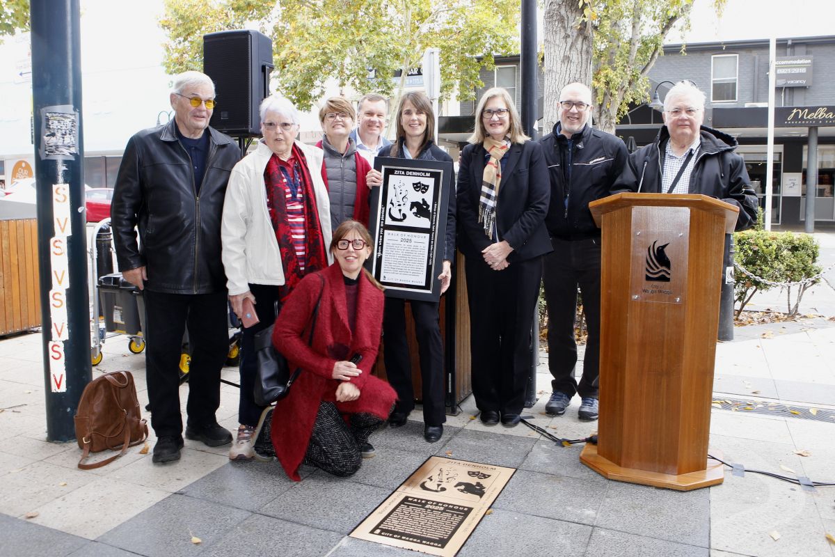 A group of people stand outside near a bronze plaque set into the tiles of a pedestrian pathway. One woman in the group holds a framed copy of the plaque.