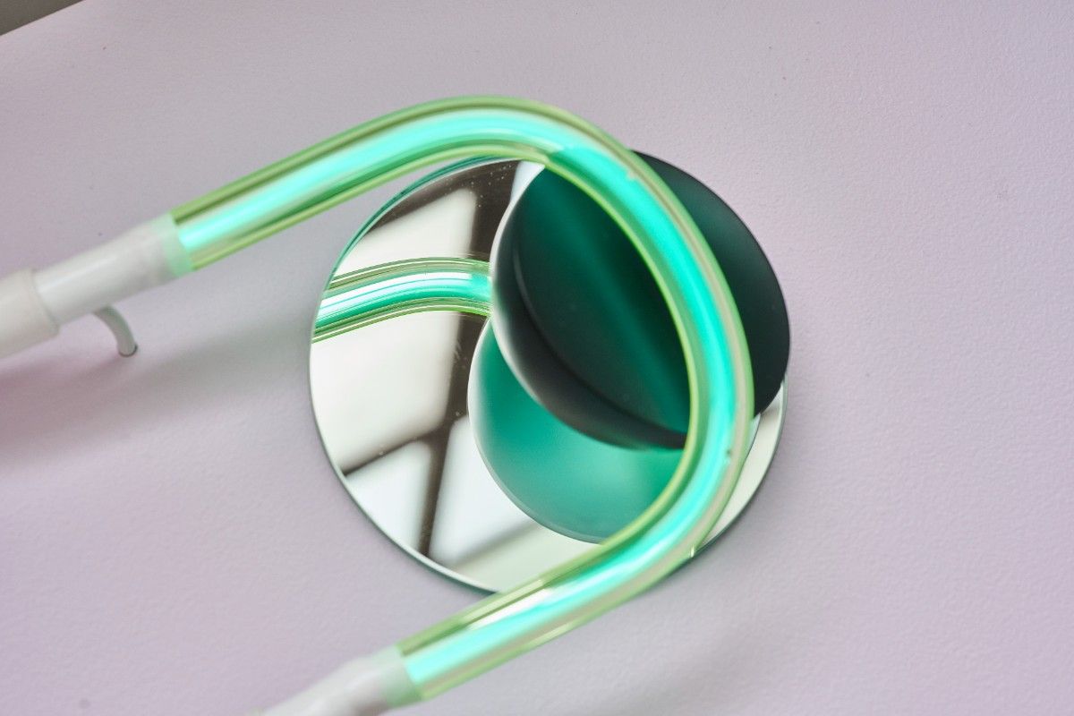 A green halogen light tube curves across a dark glass 3D shape sitting on top of a round mirror.