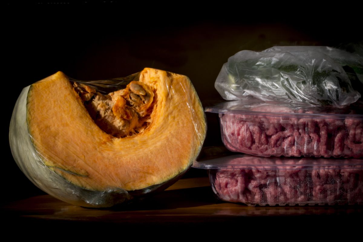 A still life photo of a pumpkin wedge, parcel of green vegetables and two plastic trays of minced meat, all encased in plastic