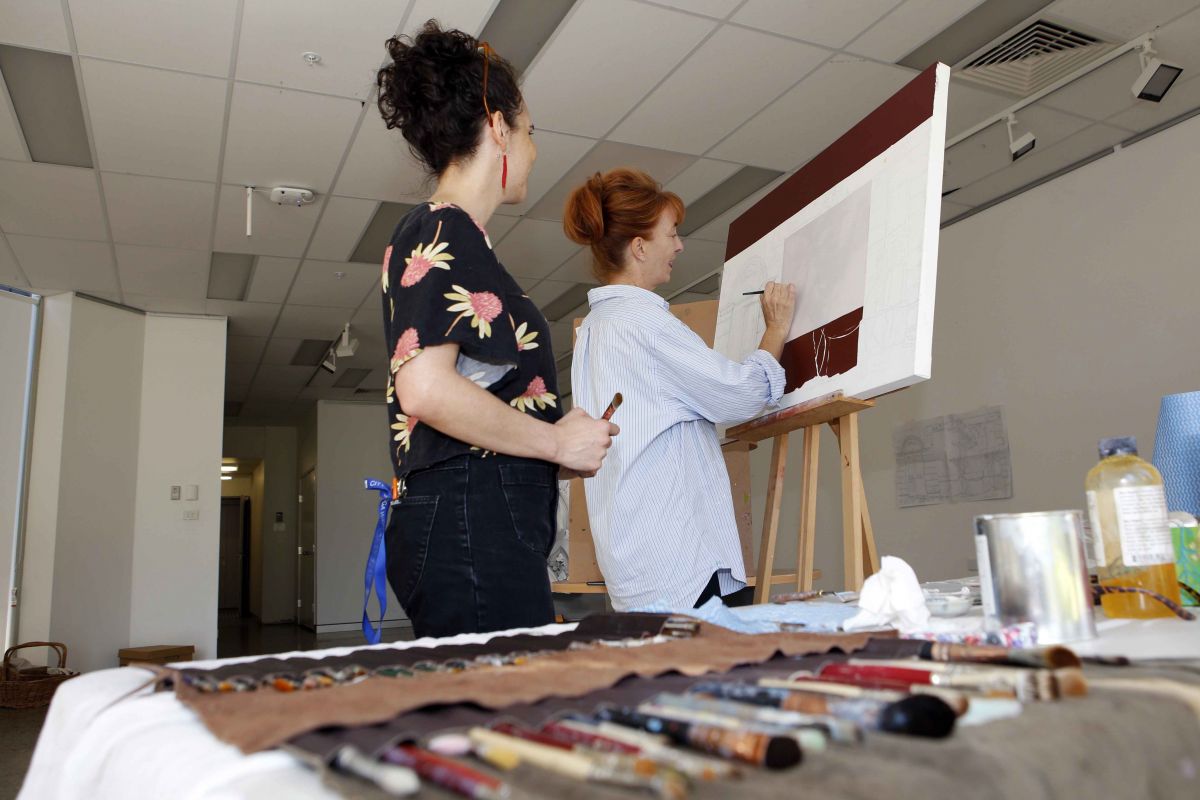 Two women standing in front of artwork with table covered in paint brushes in foreground