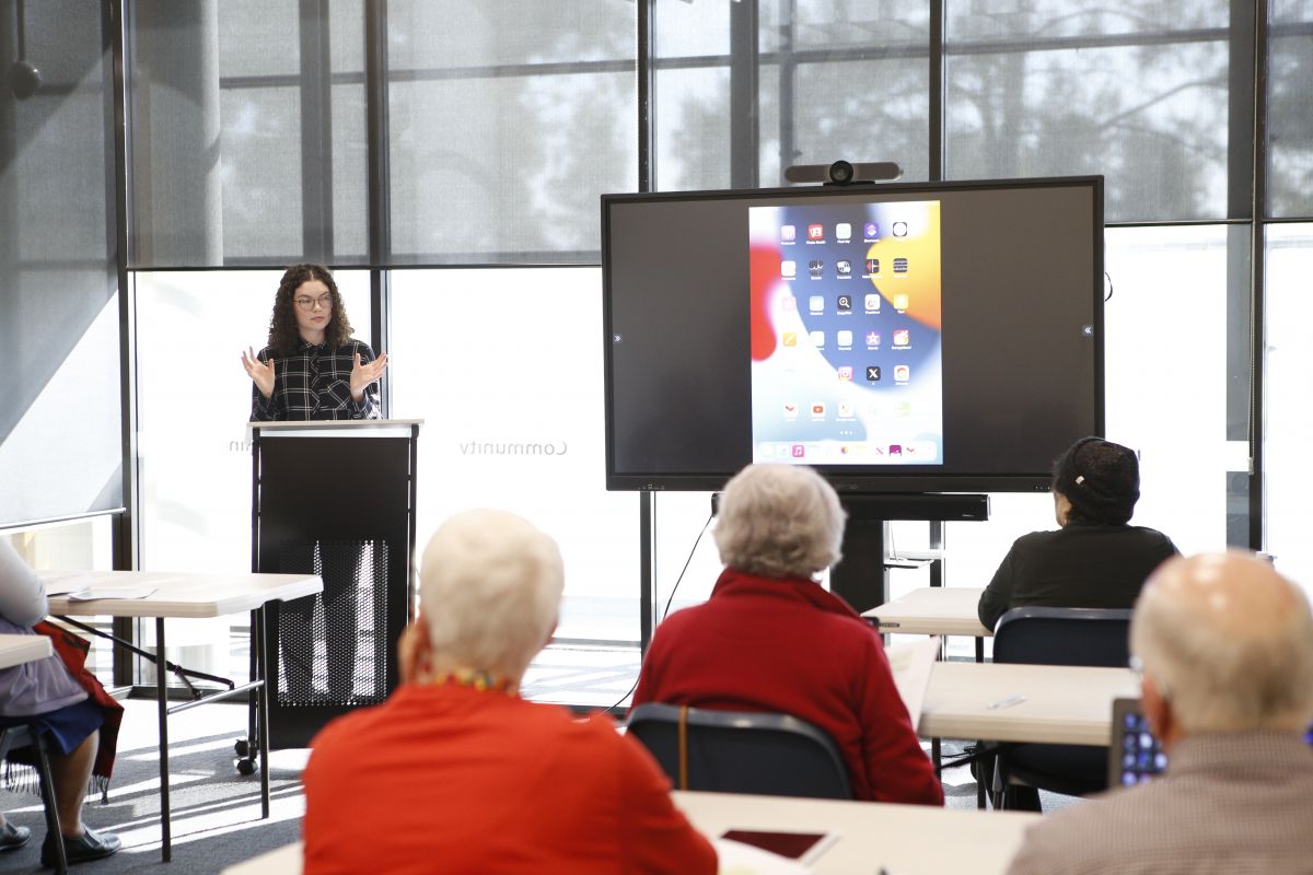 Wide shot as class trainer Sophia talks to a group of seniors and gestures to a display TV where she demonstrates how to create an email address.