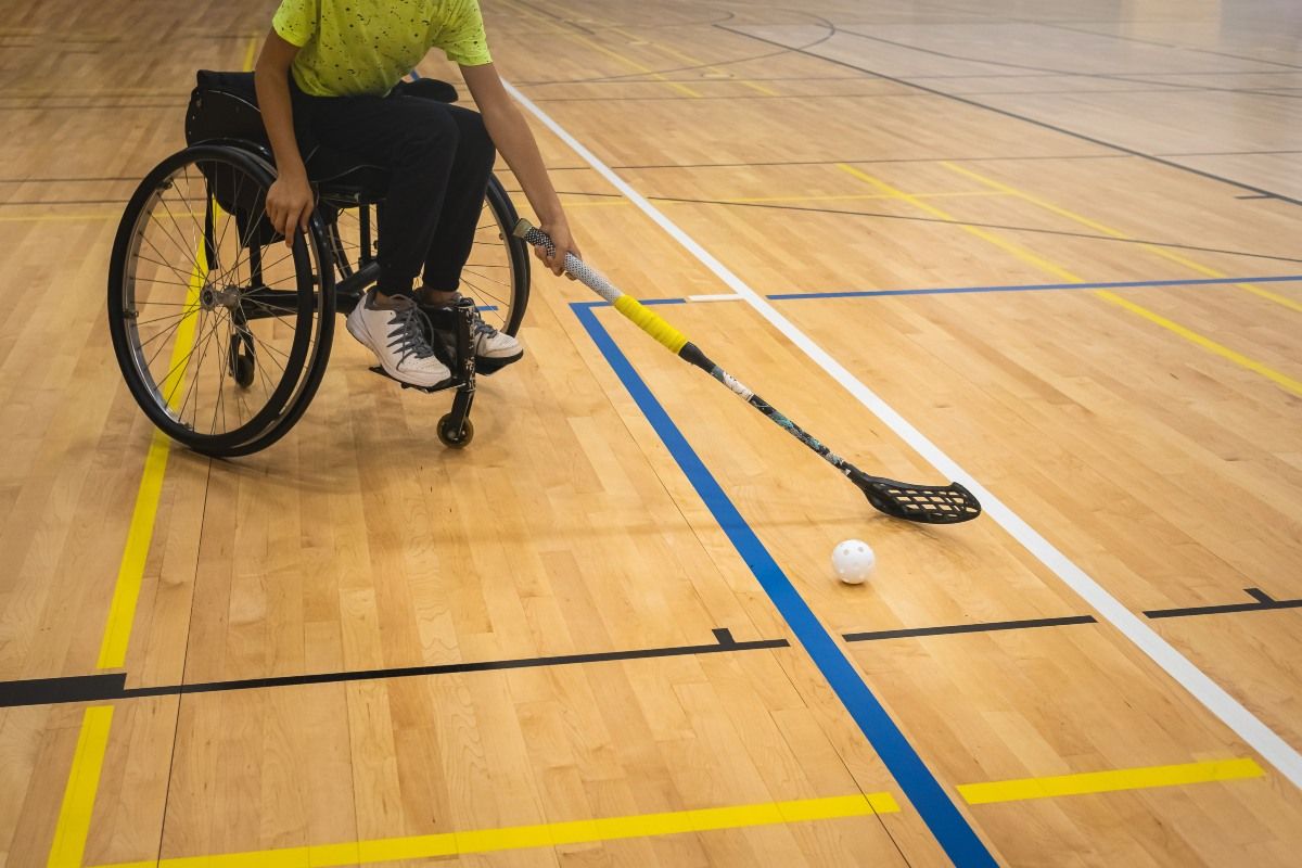 A young person in a wheelchair, on an indoor sports wooden surface. The young person is holding a lacrosse stick and there is a white ball on the floor in front of them. 