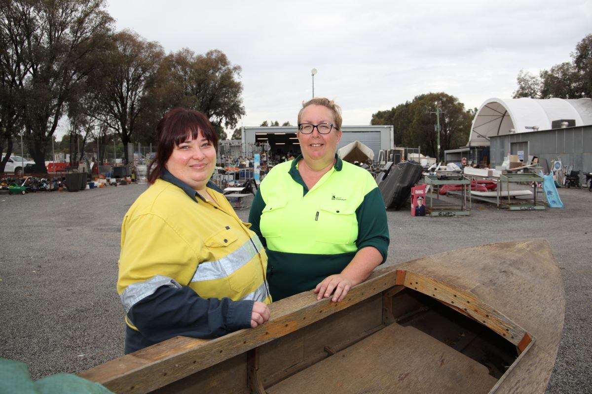 Two women wearing high vis stand in a yard with second-hand items. 