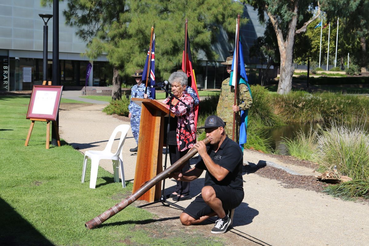 An elder Aboriginal woman speaking at a lectern, a younger Aboriginal man plays the Didgeridoo beside her. 