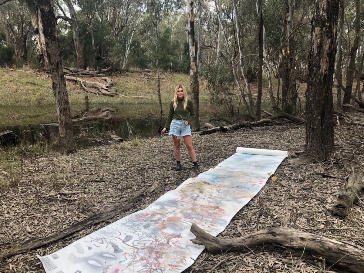 A young woman stands in a bushland with an artwork laid at her feet