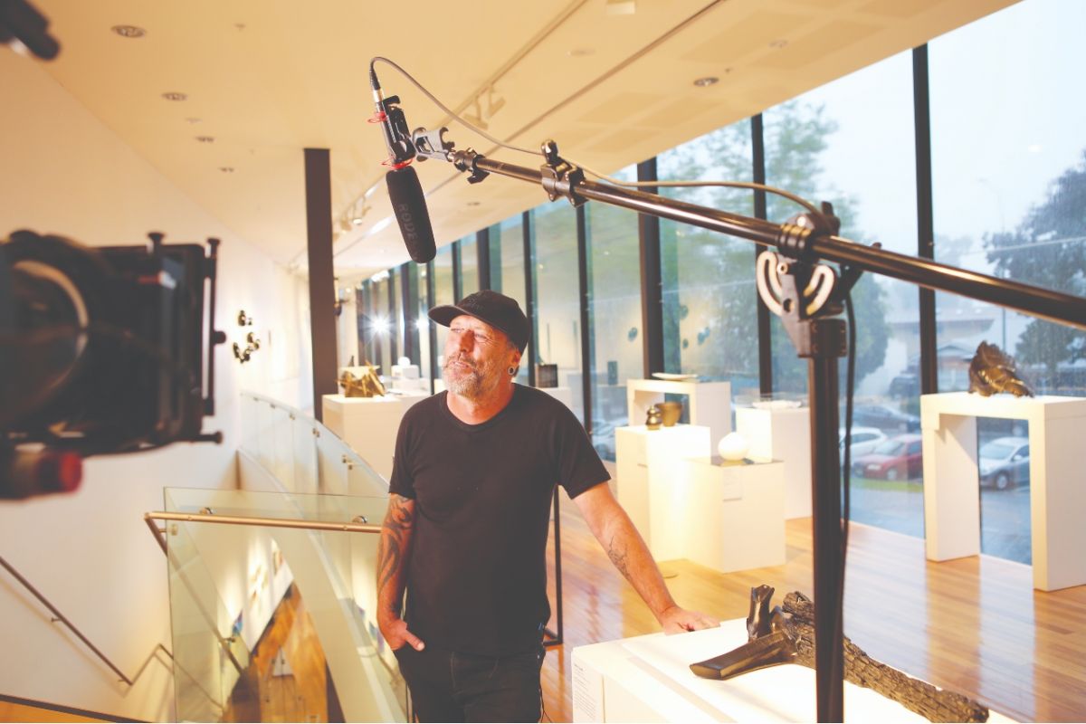 Man wearing cap standing in front of video camera, with boom mic above and art glass piece in background