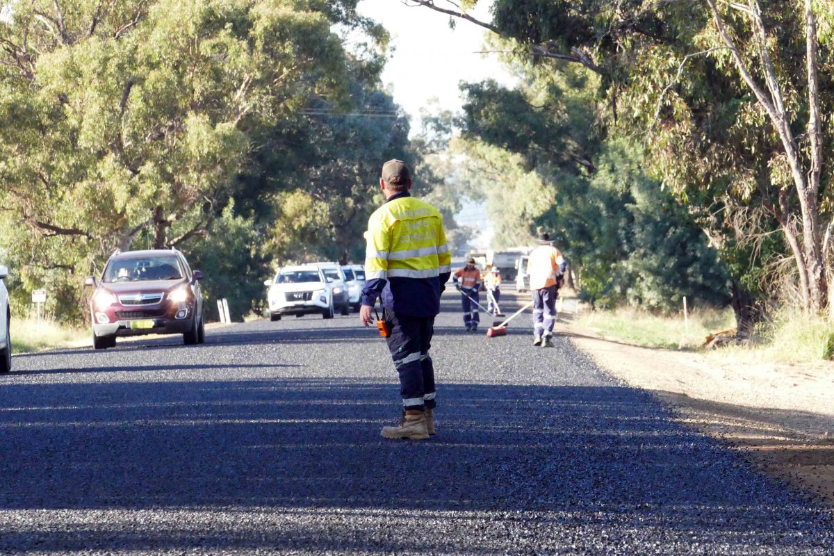 Man in hi-vis vest standing on freshly sealed road, traffic going by, workers sweeping road in background