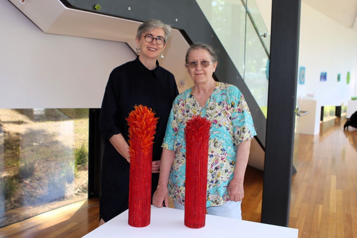 Two women in an art gallery standing with some tall red glass pieces. 