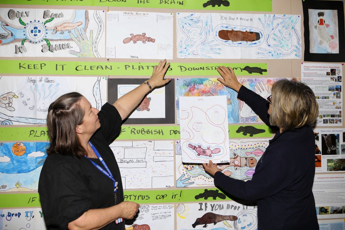 Two women stand in front of a wall of artwork, pointing and looking at the art.