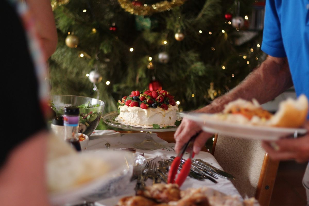 A hand holding tongs pick up a piece of food from table to place on a plate being held in the other hand. In the background is a pavlova covered in fruit, and a decorated Christmas tree.