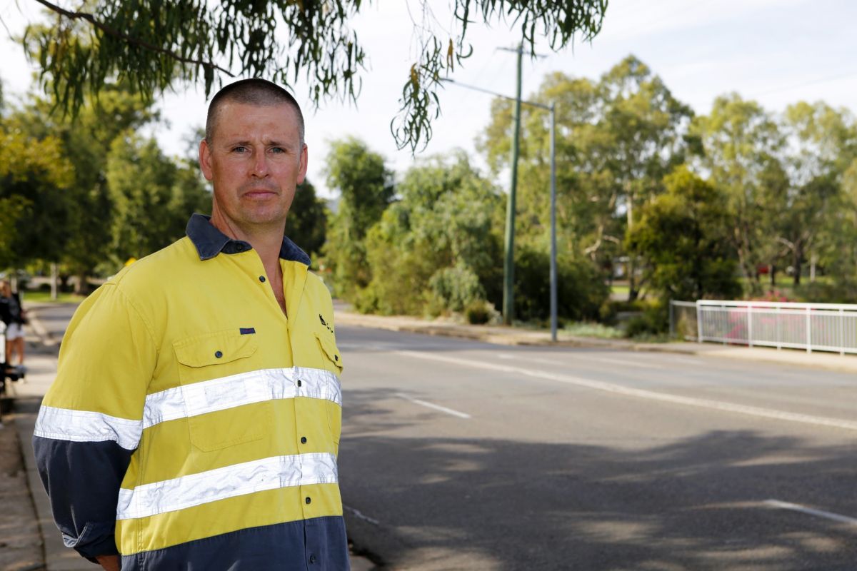 Man in hi-vis shirt standing beside a road, with a footpath and bridge railings in the background