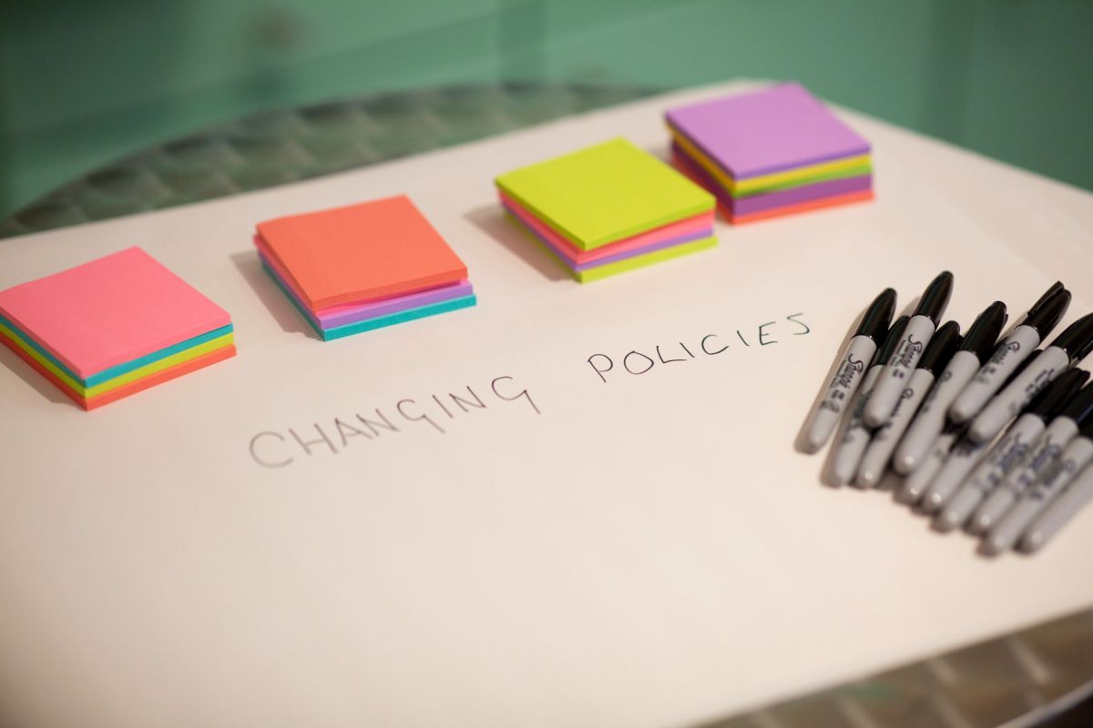 Piles of different coloured stick notes sitting on a large piece of paper, with a pile of black marker pens nearby and the words ‘changing policies’ on the paper.