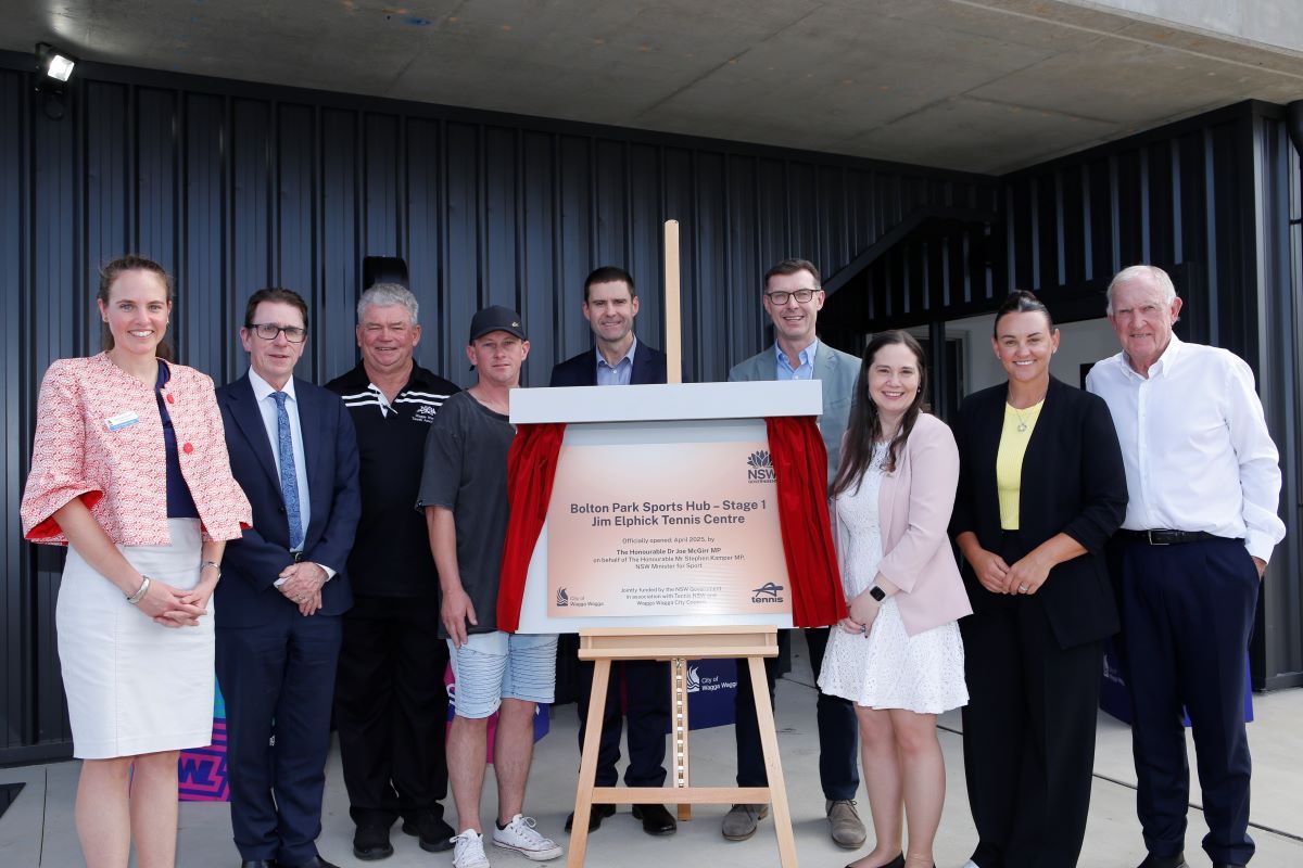 Nine people stand side by side, beside and behind a newly revealed replica bronze plaque that is flanked by two short length red velvet curtains. Six of the nine people are men, and three are women.