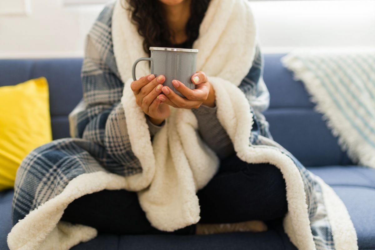 Female sitting on couch, holding hot drink, blanket wrapped around shoulders,