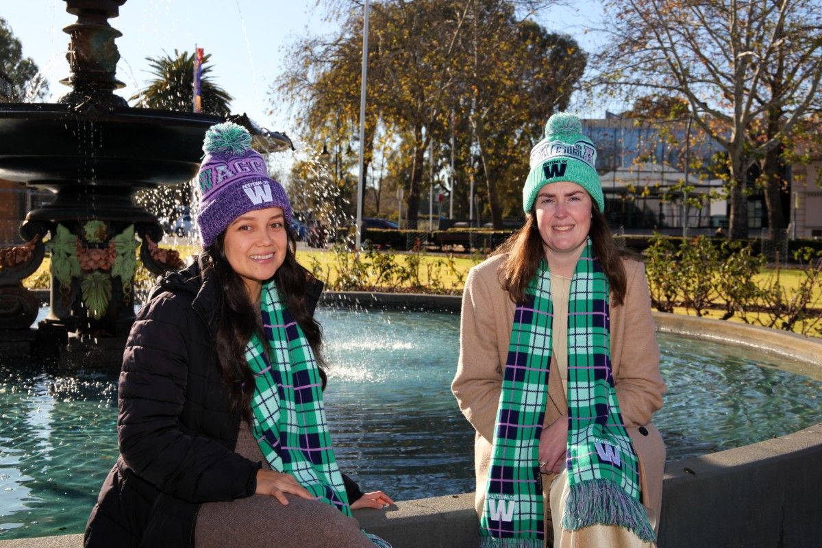 Two women sitting on the edge of a large fountain, with both women wearing colourful beanies with Festival of W stitched on front, along with green and purple tartan scarves.