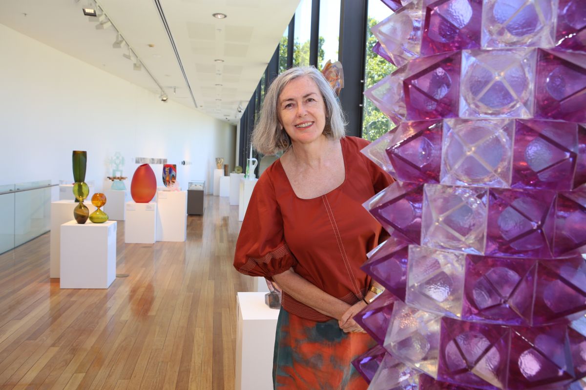 Woman standing next to large glass art piece with other glass art works in background