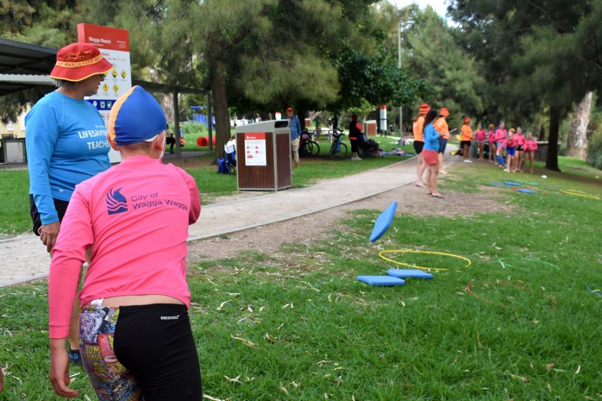 Child in pink shirt throws a kick board towards a ring in a park