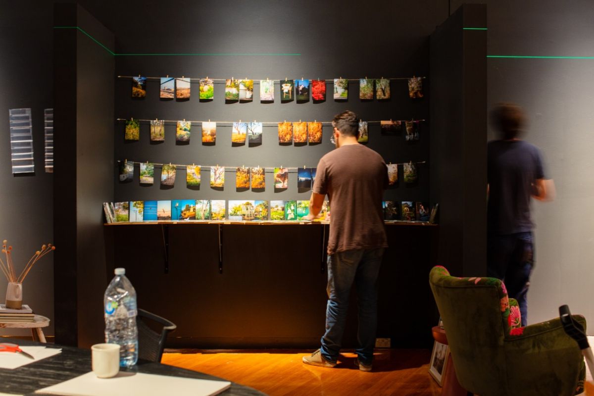 A man stands with his back facing the viewer in front of a black wall hanging a series of photographs on a string line.