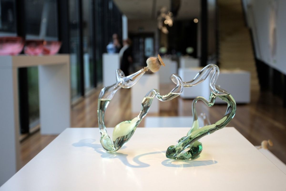 A poppy pod sits in an artistically formed glass vase filled with pale green liquid