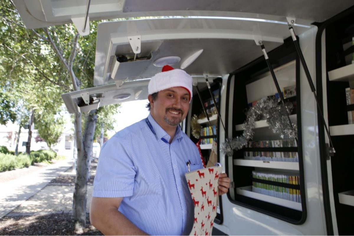 A smiling man in a Santa hat holding a Christmas stocking beside a van filled with books