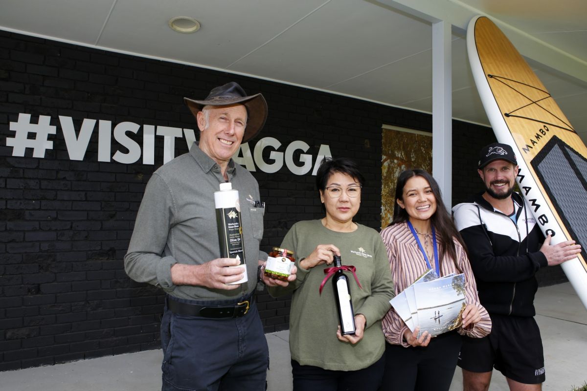 Two women & two men, one holding a stand-up paddle board, stand outside Visitor Information Centre with #visitwagga sign in background