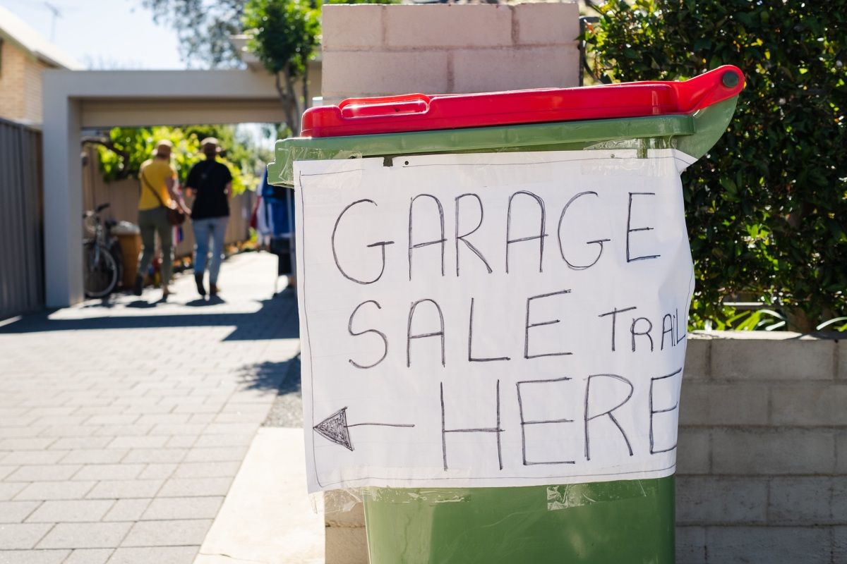 Close up image of a red wheelie bin with a sign taped to its side. The sign reads 'Garage Sale Trail here' with an arrow drawn to point to the left. 