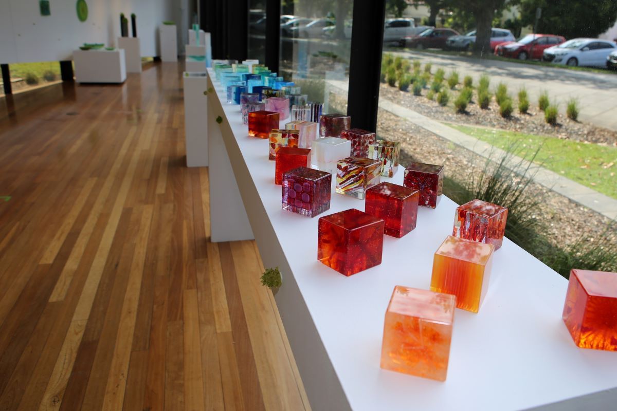 small glass cubes of various colours on a long table in a gallery in front of a window.