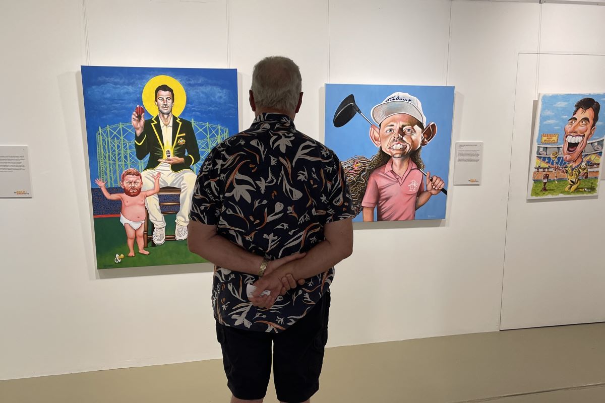 The back of a man standing in an art gallery space looking at artworks. The artworks on the wall are caricature portrait paintings of Australian sporting personalities. 