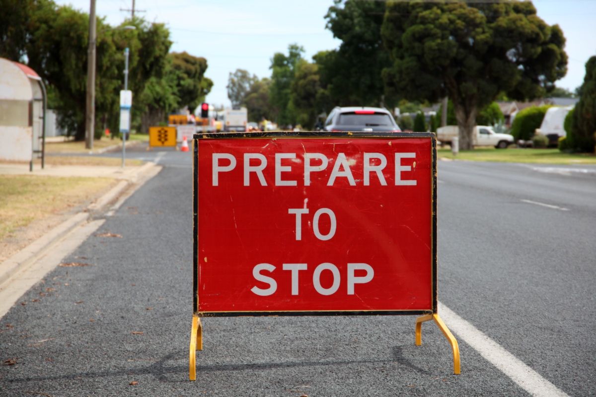 Red roadworks sign saying ‘prepare to stop’ on side of the road, with traffic control, vehicles and roadworks in the background.