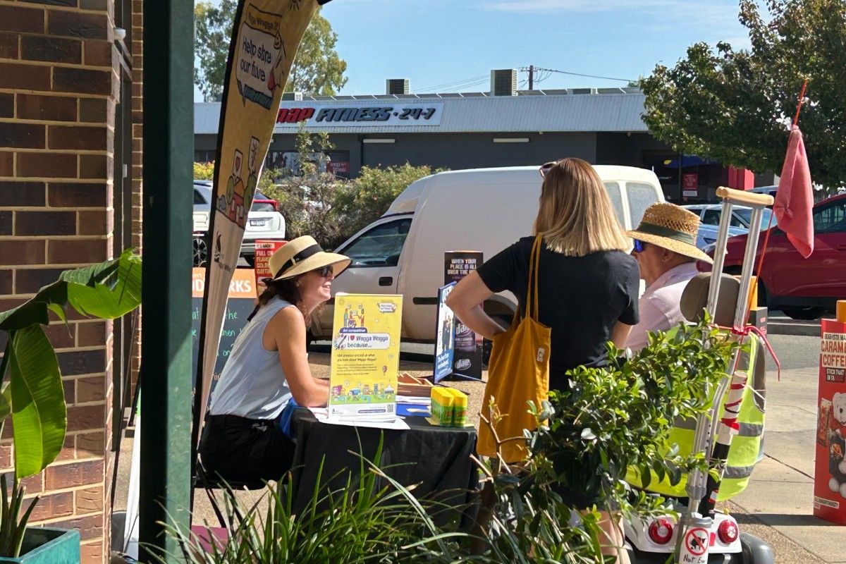 Woman wearing a hat sitting at a table outside a brick building, talking to two people, including a man in a mobility vehicle.