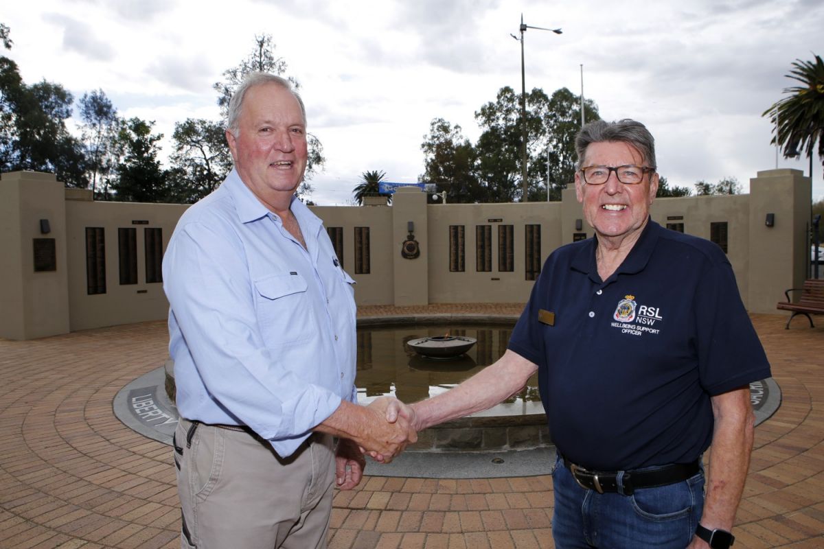 Two men shaking hands, with an Eternal Flame in a pond and Honour Rolls on the walls of a memorial in the background.