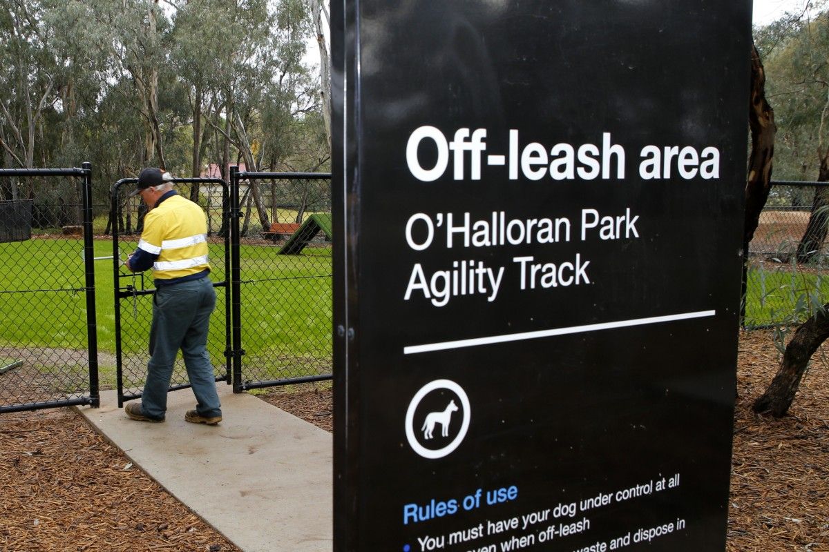 Sign saying Off-leash area, O'Halloran Park Agility Track in the foreground with a man in a hi-visibility shirt in the background opening a chain-link gate.