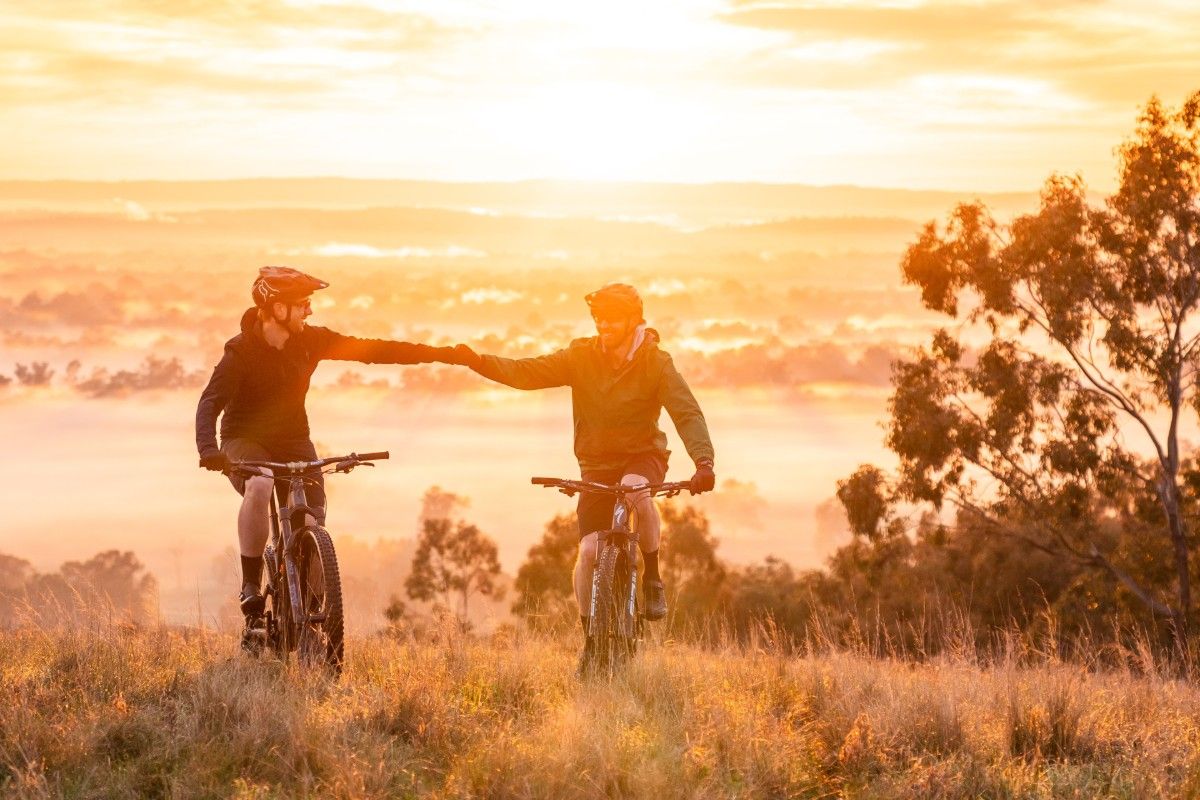 Two men wearing cycling helmets give each other a fist bump as they ride the mountain bikes up a hill.