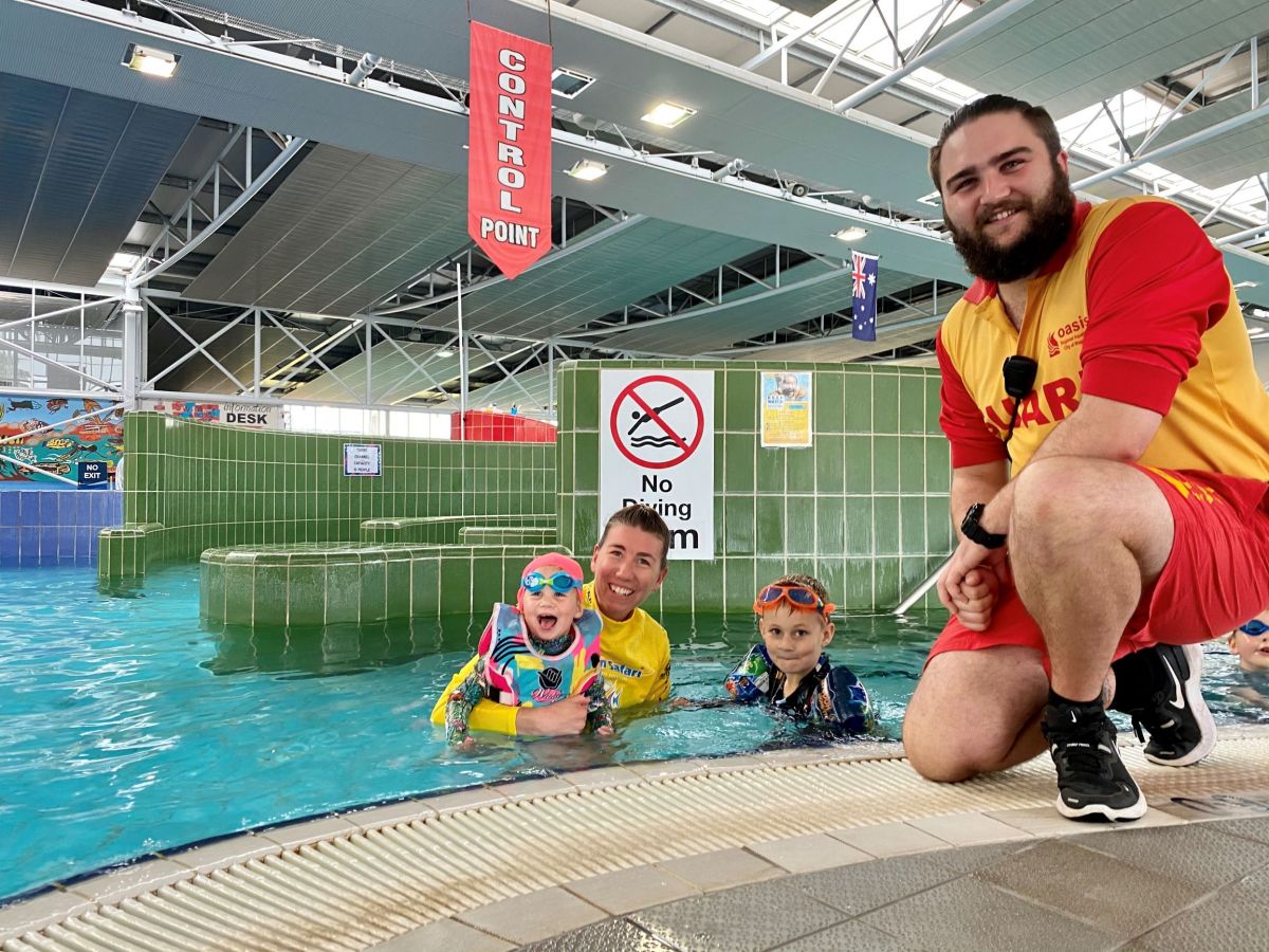 A woman with two young children in a public pool, beside a lifeguard