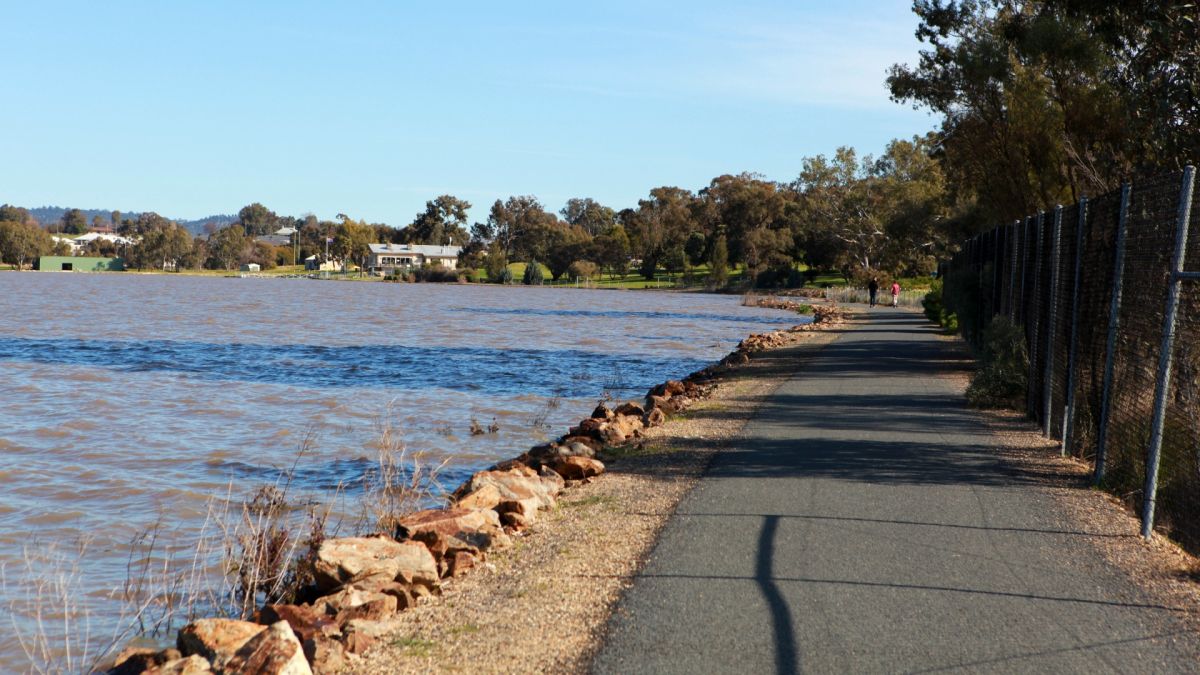 Path along banks of Lake Albert with two people walking and Boat Club in distance