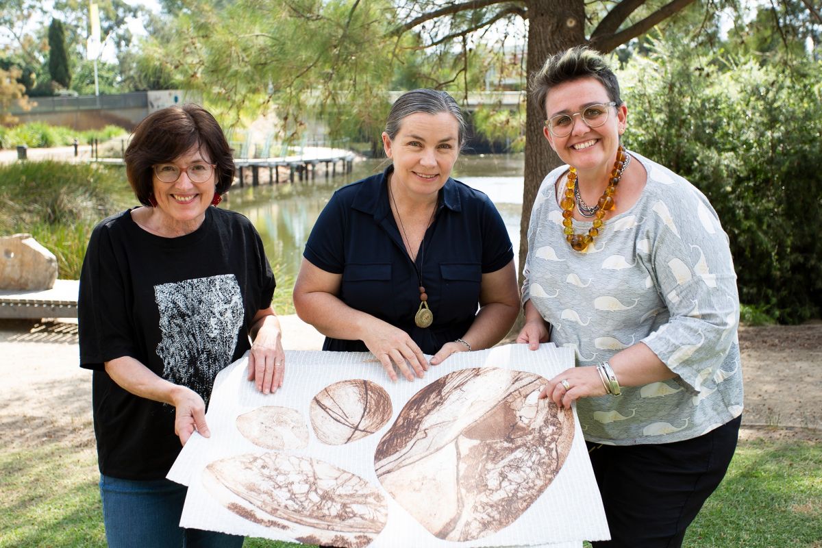 Three women in a parkland display an artwork