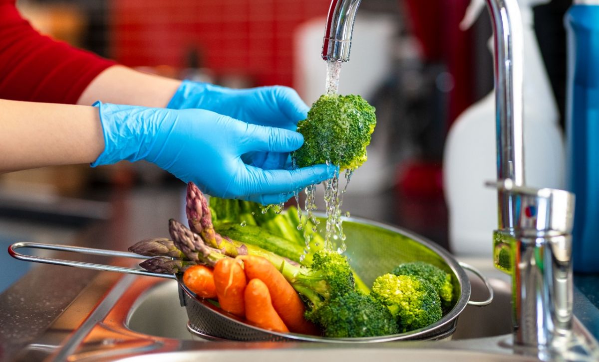 Women wearing blue food handling gloves washing vegetables under running water from tap